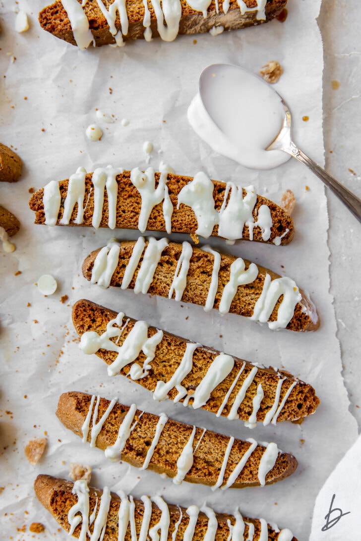 Gingerbread biscotti on wax paper with white chocolate drizzle and spoon of melted white chocolate