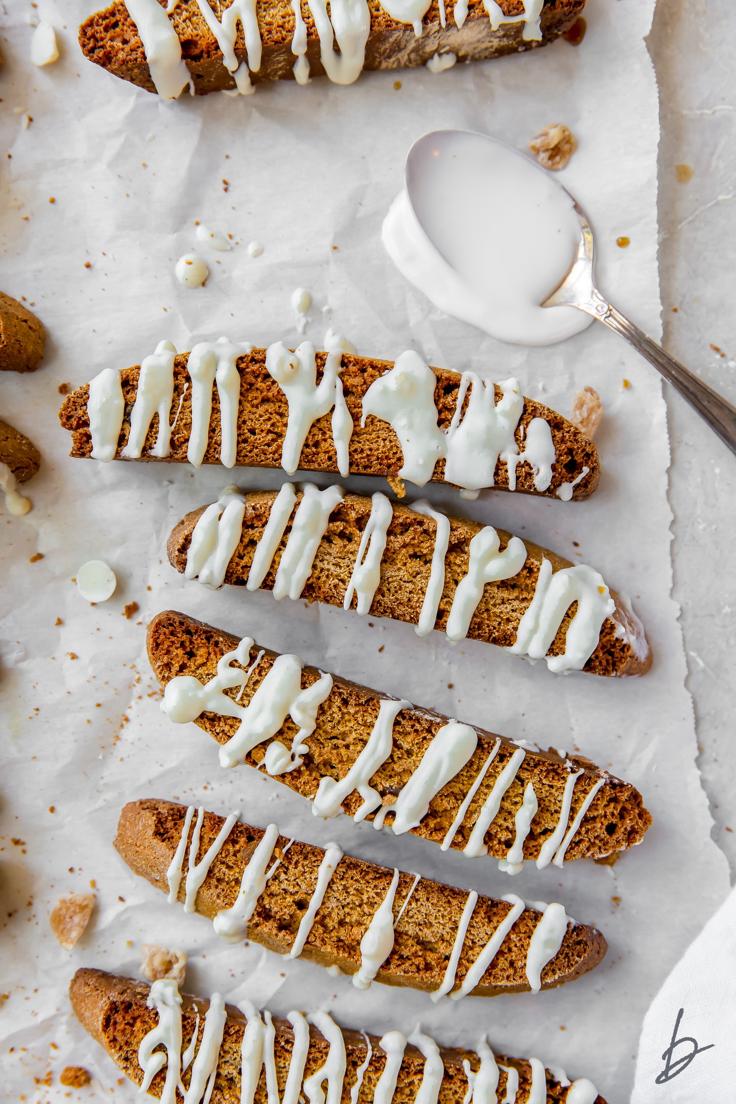 Gingerbread biscotti on wax paper with white chocolate drizzle and spoon of melted white chocolate.