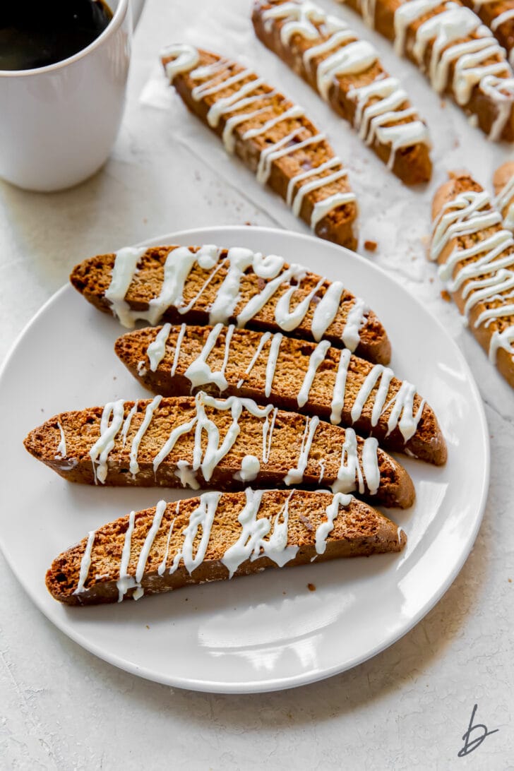 Gingerbread biscotti with white chocolate drizzle on white round plate next to white mug of coffee