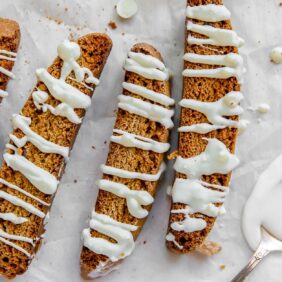 gingerbread biscotti drizzled in white chocolate on top of parchment paper with spoon of melted white chocolate