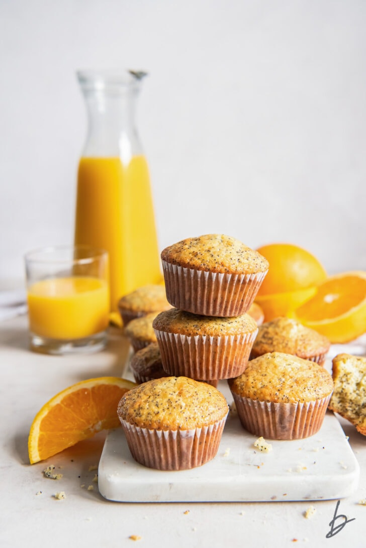 orange poppy seed muffins stacked on top of each other in front of pitcher of orange juice and fresh oranges
