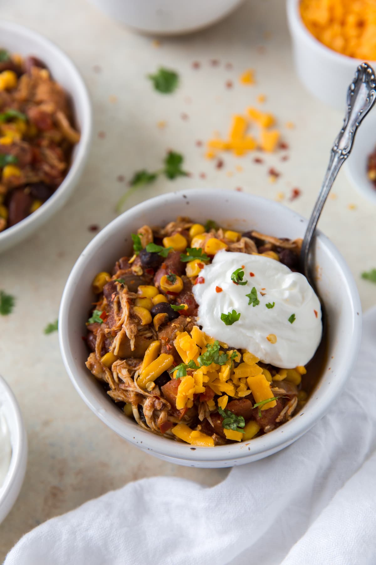 chicken chili topped with shredded cheese and sour cream in a white bowl with spoon