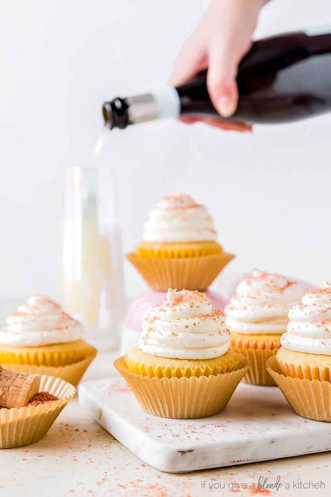 champagne cupcakes on marble slab; hand pouring bottle of champagne into glass behind cupcakes