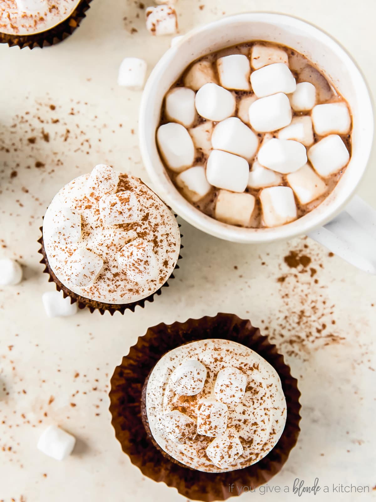 two hot cocoa cupcakes with marshmallow frosting next to mug of hot cocoa with mini marshmallows
