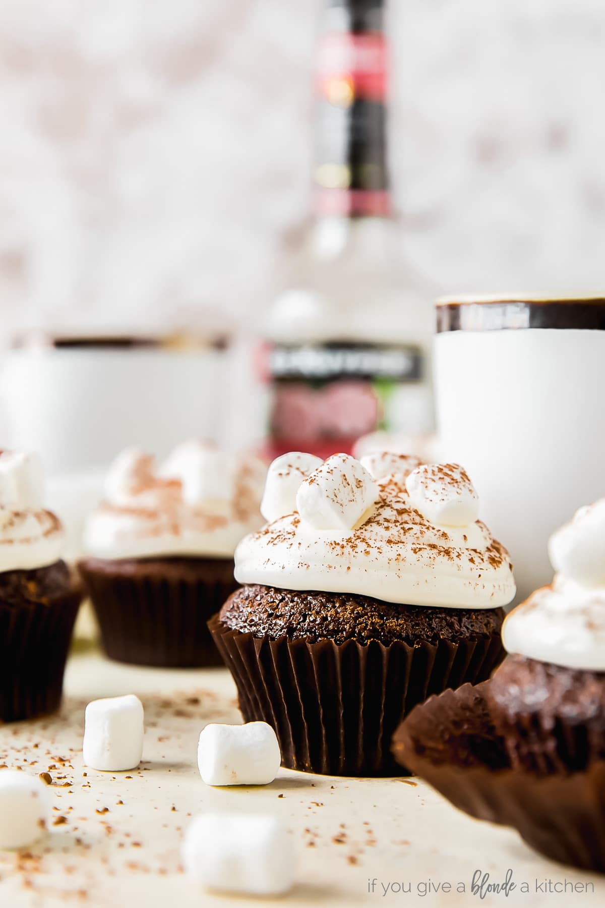 hot cocoa cupcake topped with marshmallow frosting, mug behind cupcake