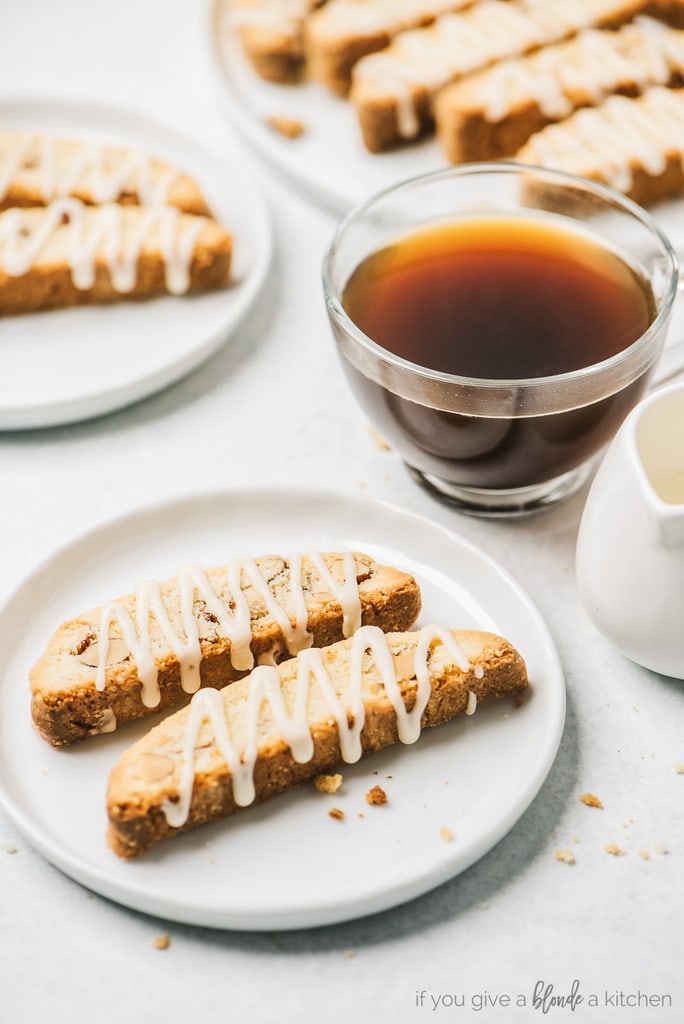 two almond biscotti with vanilla glaze on round white plate; clear mug of coffee next to plate