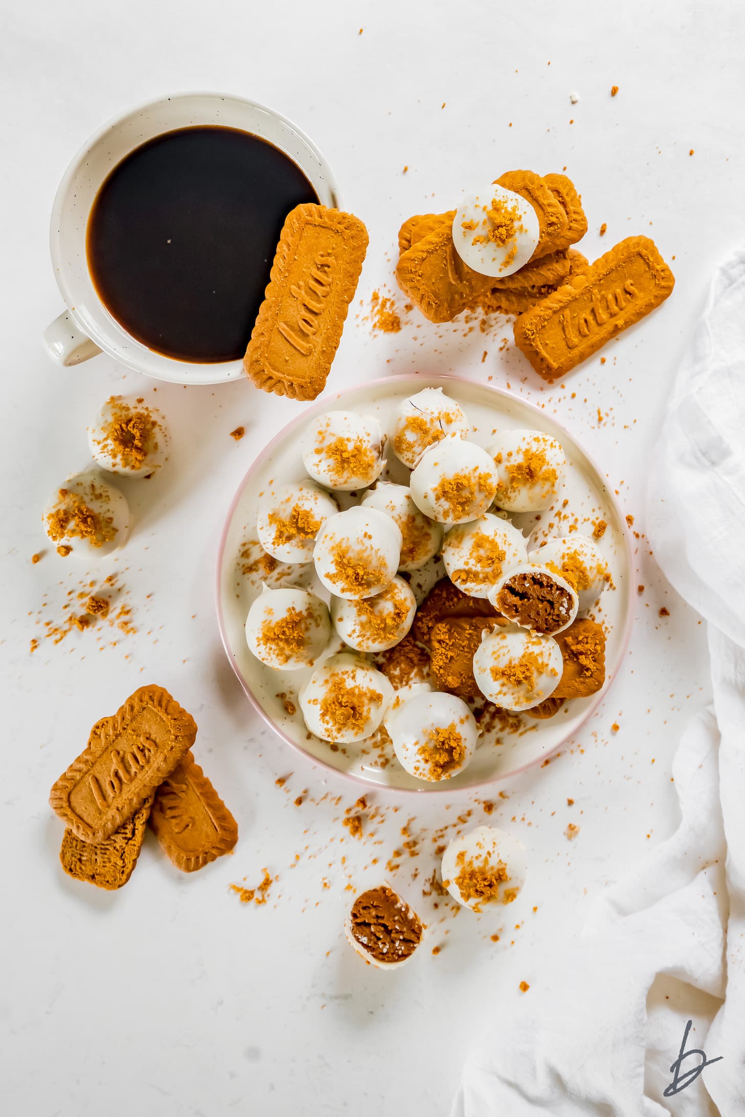 plate of white-chocolate covered biscoff truffles with cookie crumbs next to more biscoff cookies and mug of coffee
