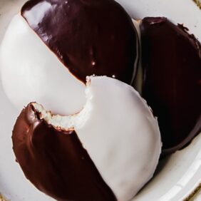 three black and white cookies on a white round plate; top cookie with a bite
