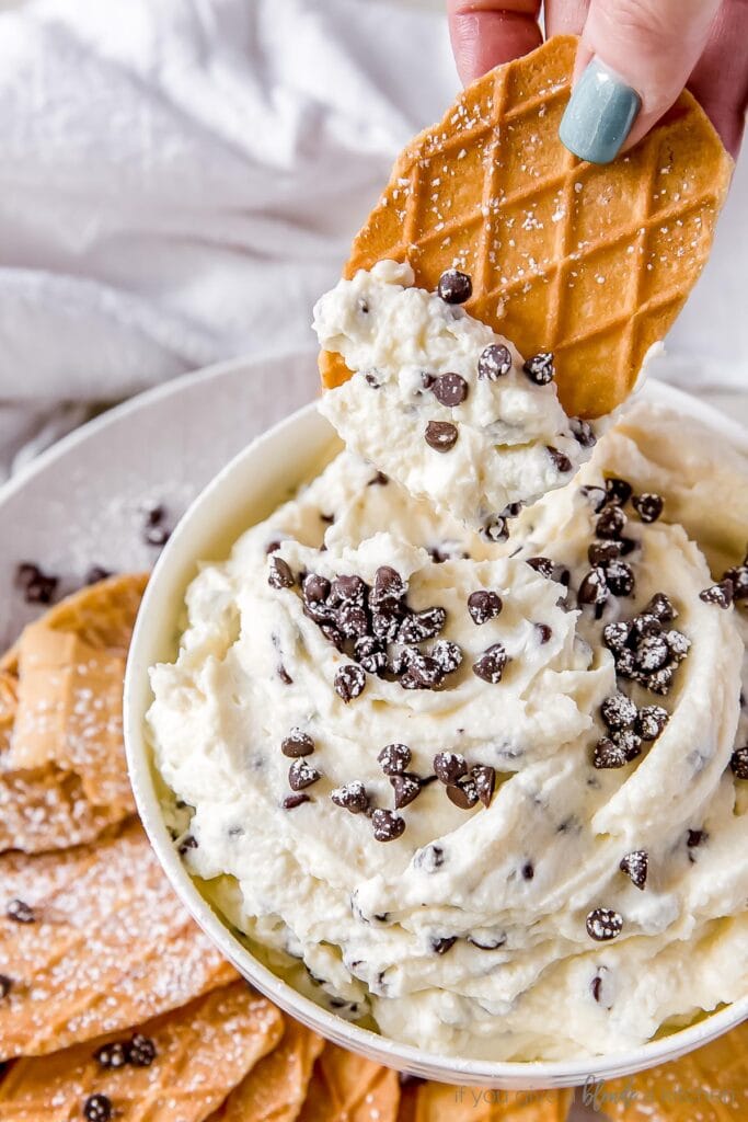 hand dipping waffle cookie in bowl of cannoli dip with mini chocolate chips