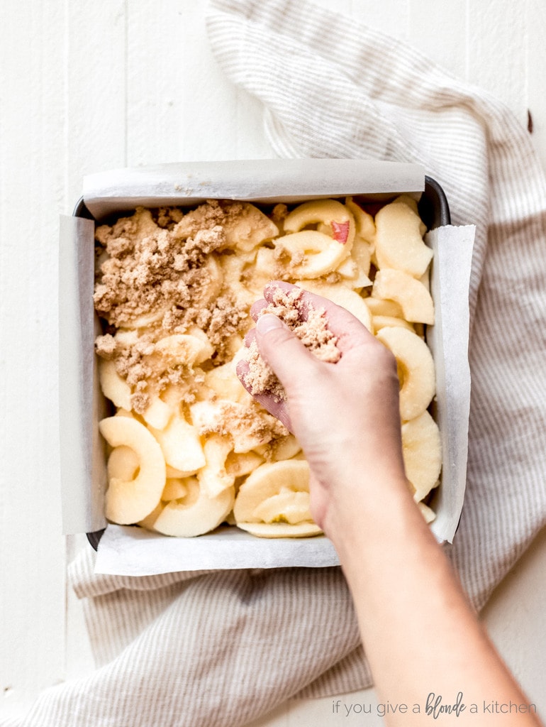 hand sprinkling crumble on top of apples in 8x8 pan