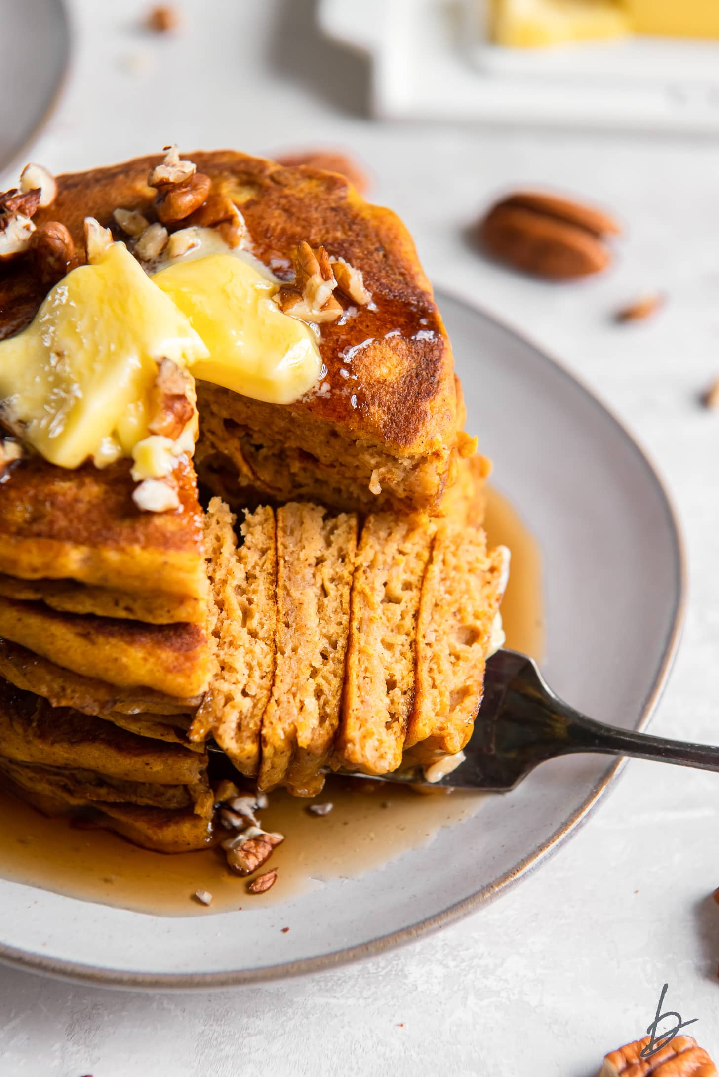 fork holding bite next to stack of pumpkin pancakes topped with butter and chopped pecans.