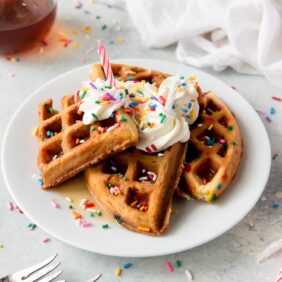 plate of birthday cake mix waffles topped with whipped cream, sprinkles and a birthday candle