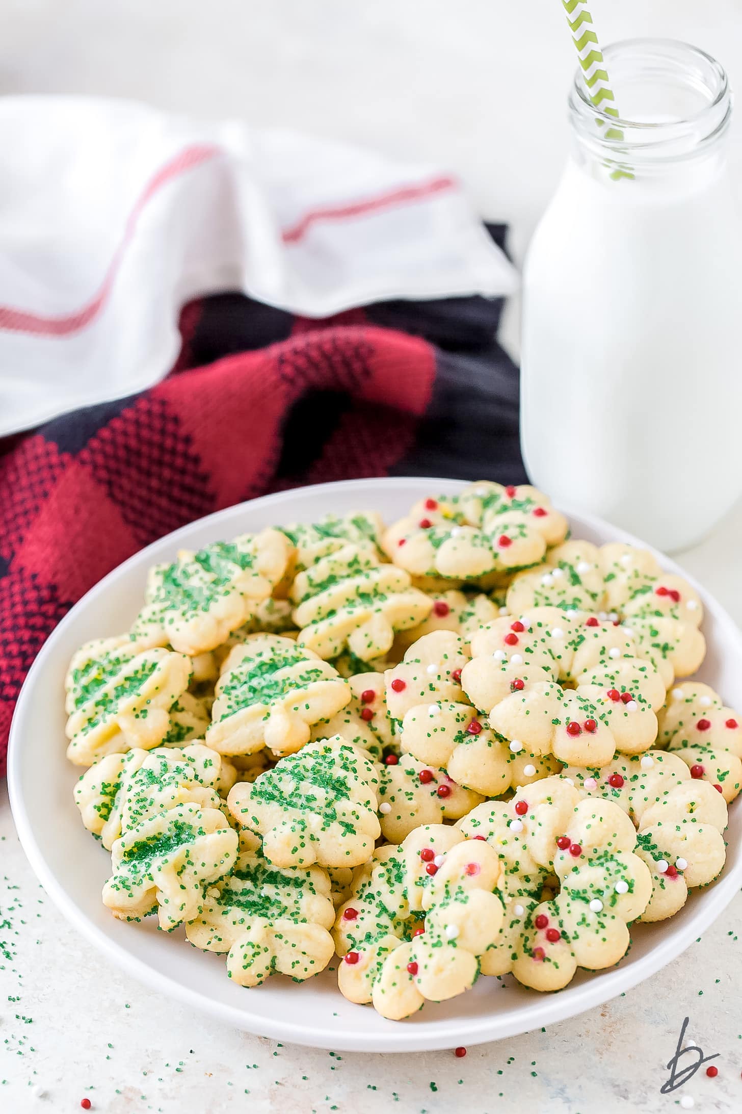 christmas spritz cookies on white plate next to bottle of milk and red kitchen towel behind plate.
