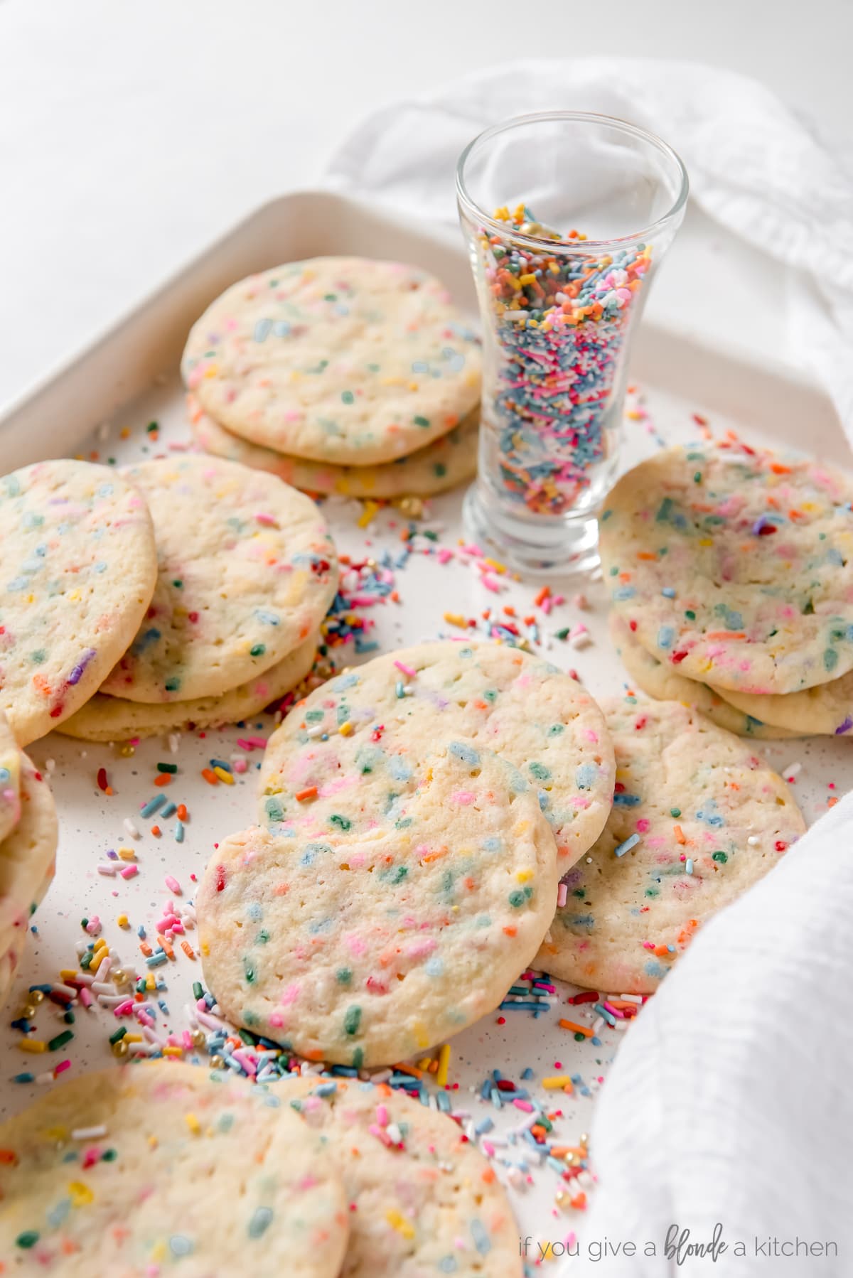 sprinkle sugar cookies piled on baking sheet with shot glass of rainbow sprinkles