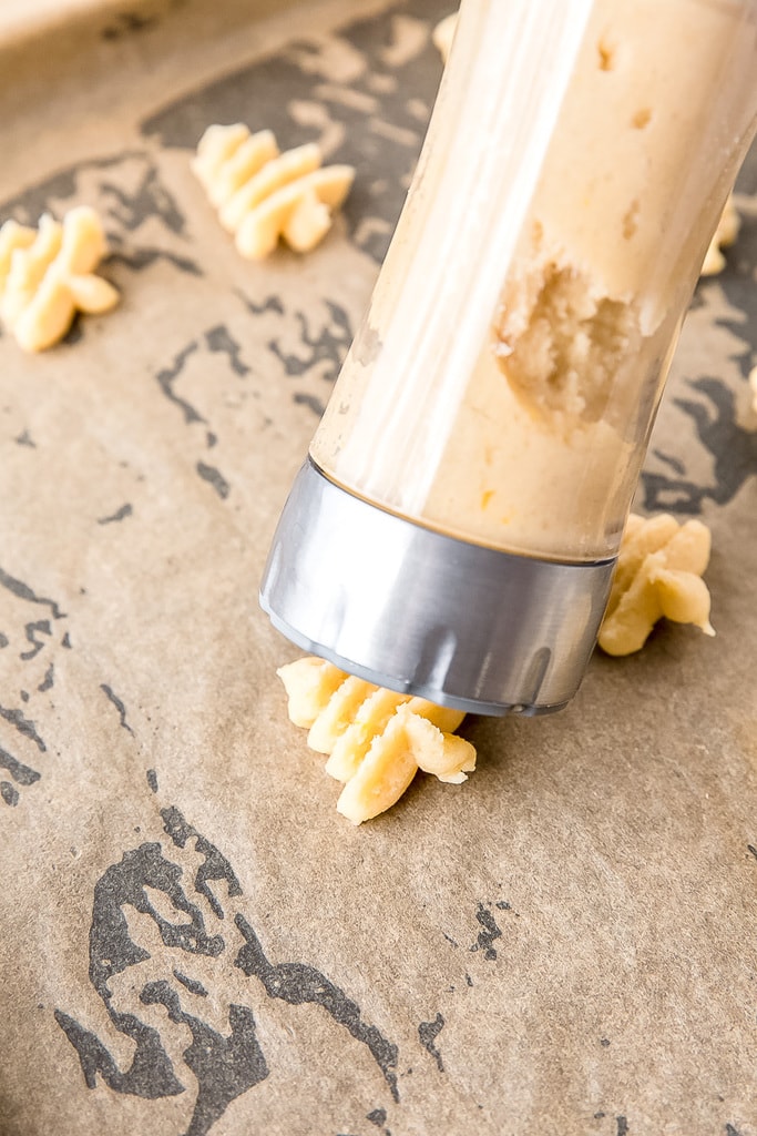 spritz cookie press pushing cookie dough into tree shape onto parchment lined baking sheet