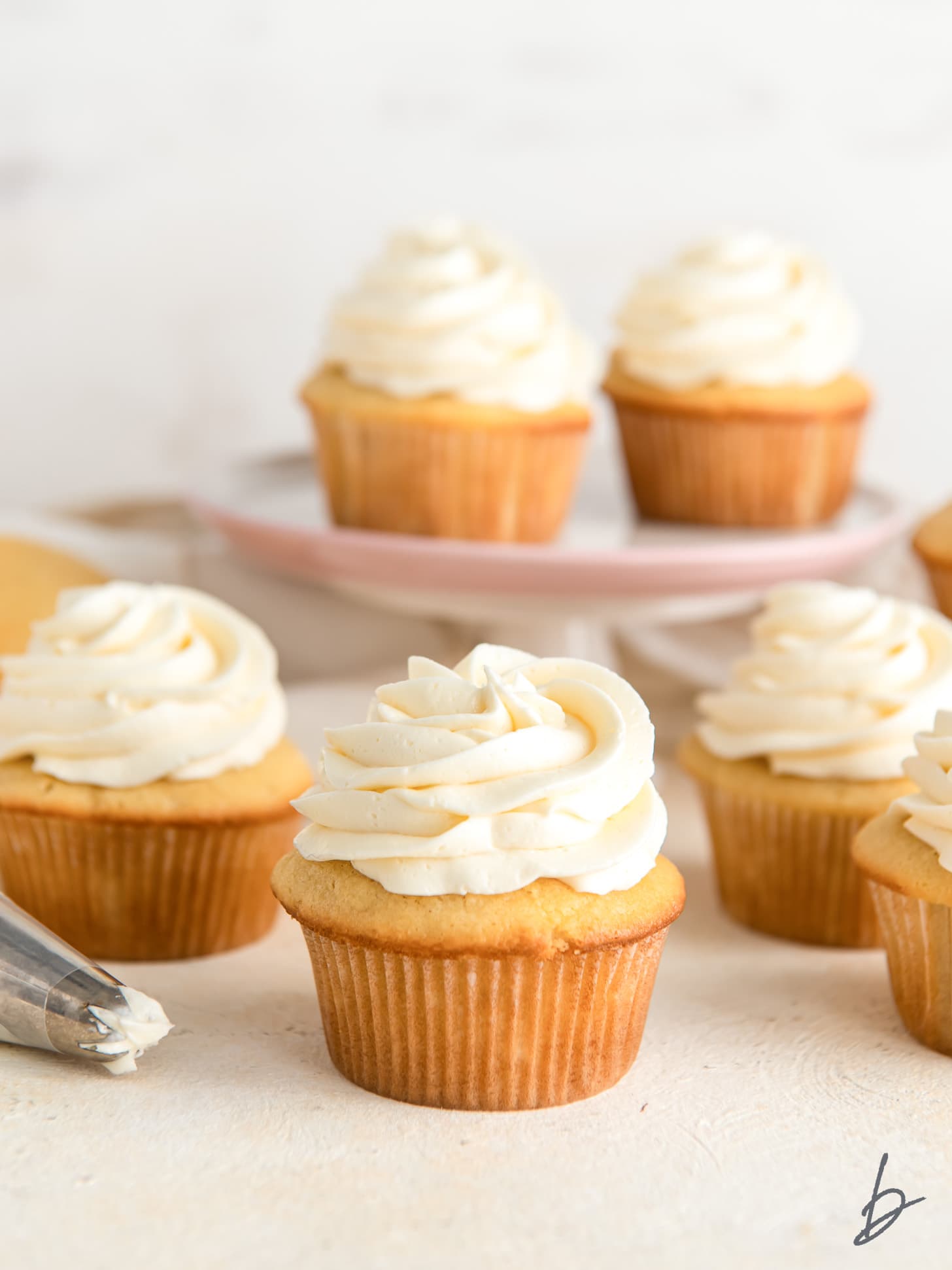 vanilla cupcakes with vanilla buttercream frosting in front of cake stand with more cupcakes