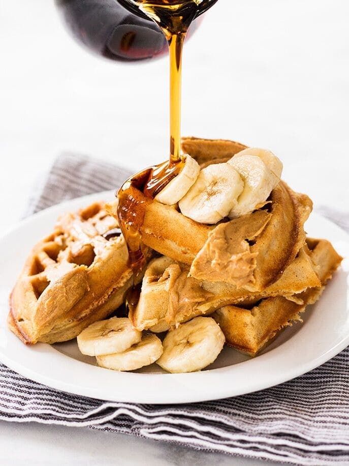 syrup poured on stack of waffles with peanut butter and banana slices