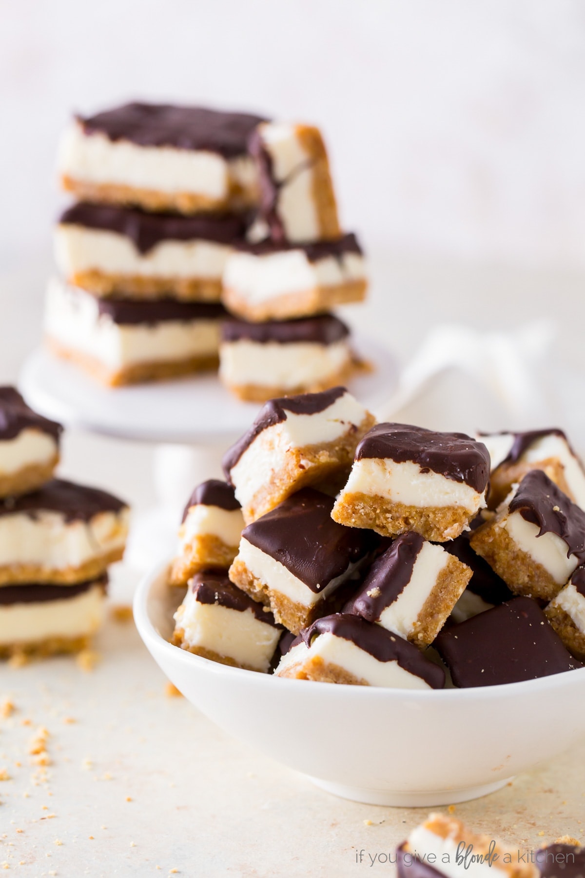 chocolate dipped cheesecake bites in white bowl with more cheesecake bites on stand behind bowl