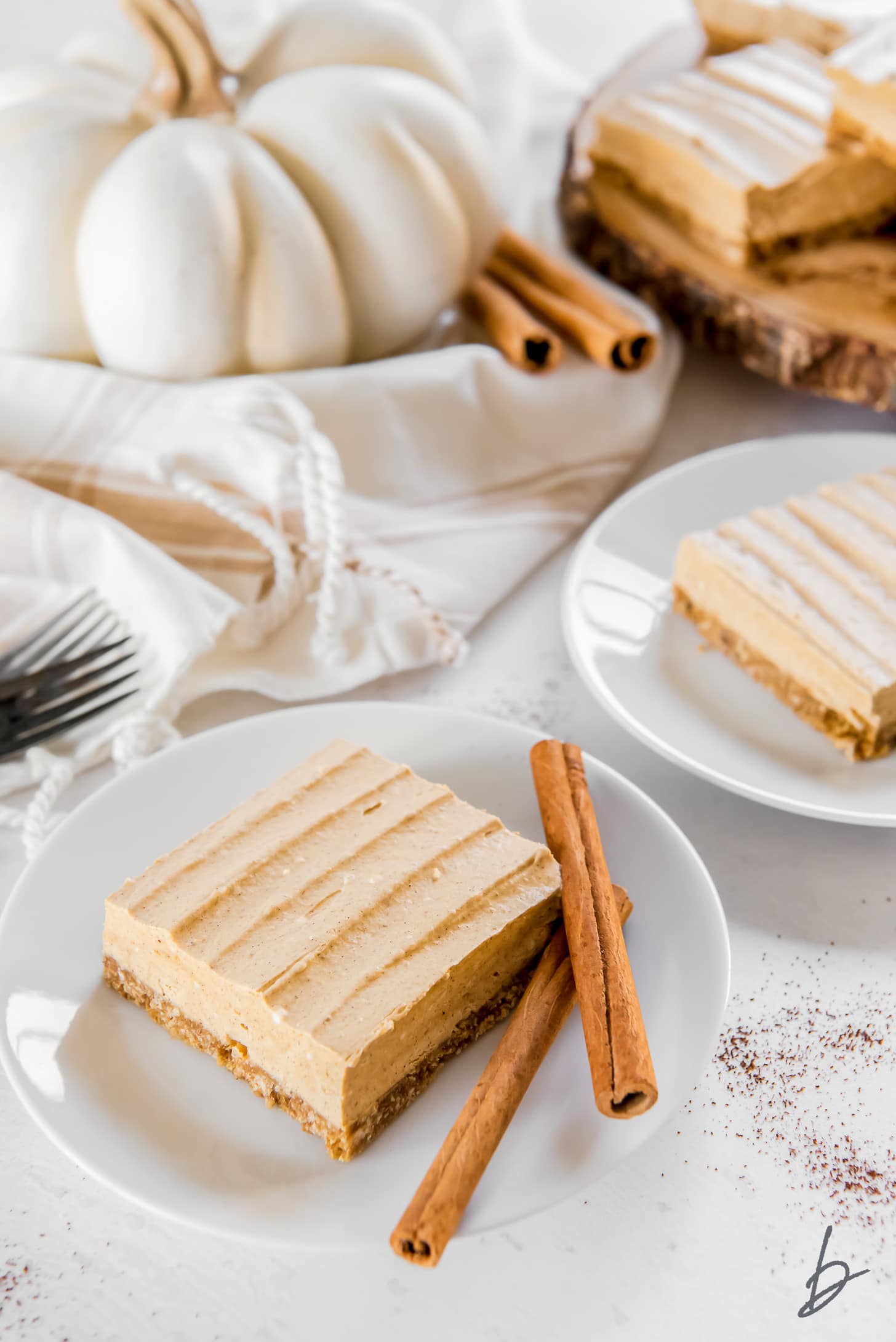 pumpkin cheesecake bar on a white round plate with two cinnamon sticks and a white pumpkin behind plate