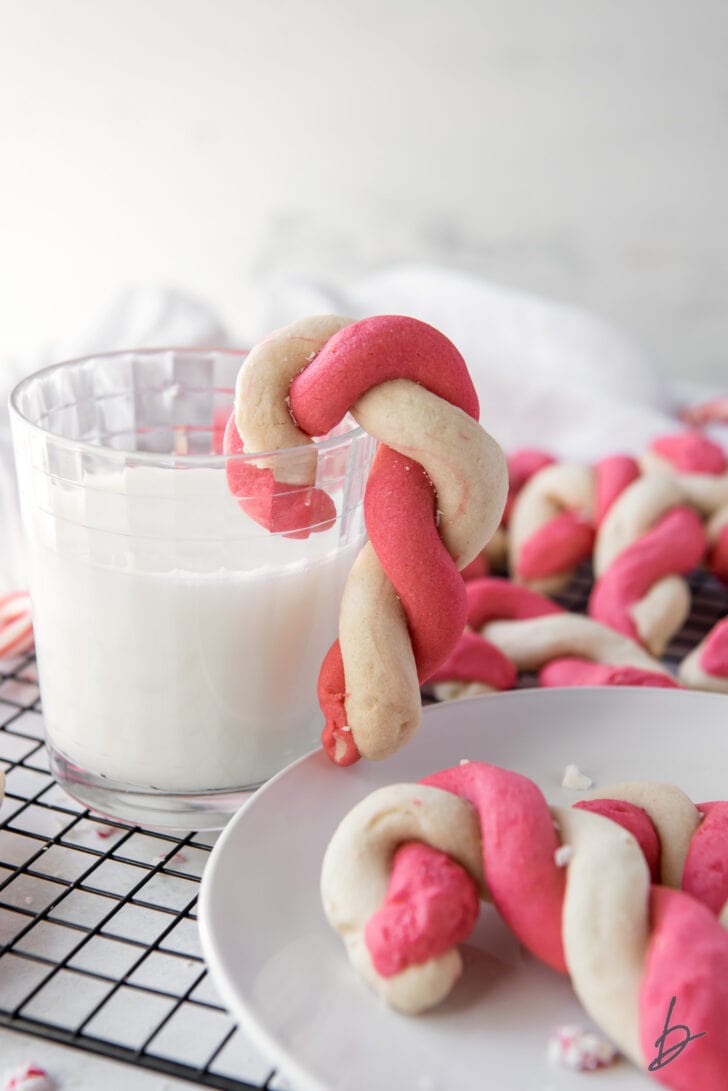 twist candy cane cookie hanging off glass of milk next to plate of cookies on wire cooling rack