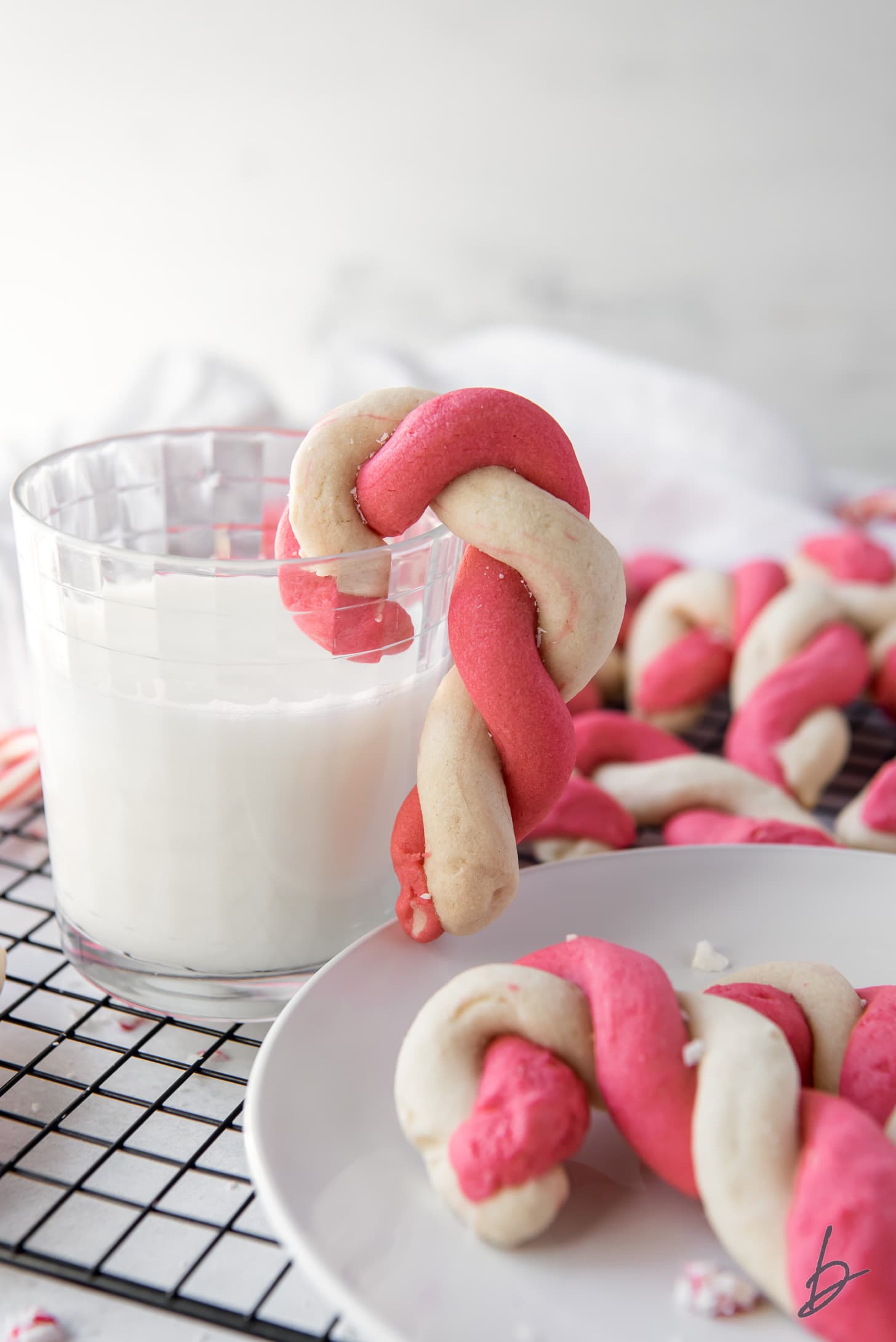 twist candy cane cookie hanging off glass of milk next to plate of cookies on wire cooling rack.