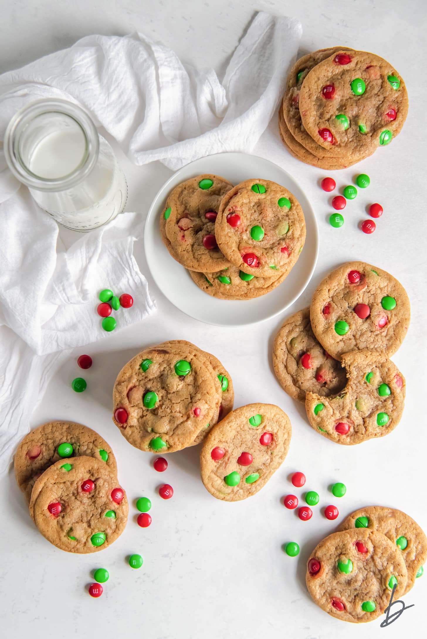 red and green M&M christmas cookies on white surface next to white kitchen cloth and glass bottle of milk