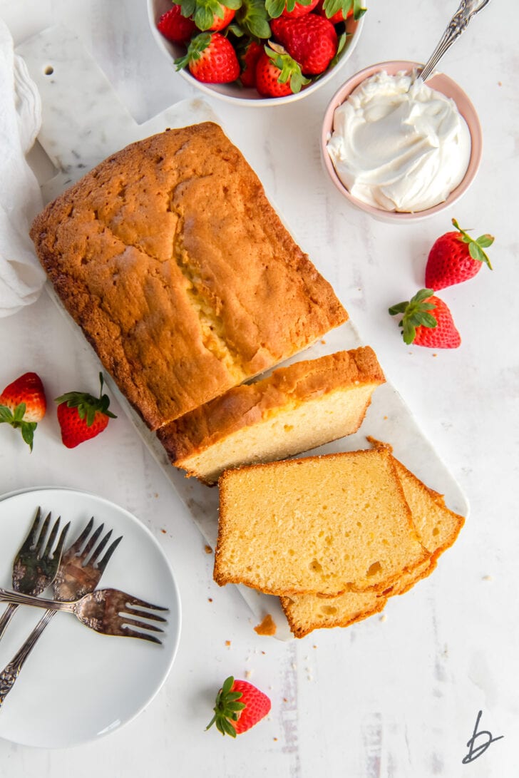 cream cheese pound cake loaf with slices cut off end next to bowl of whipped cream and fresh strawberries