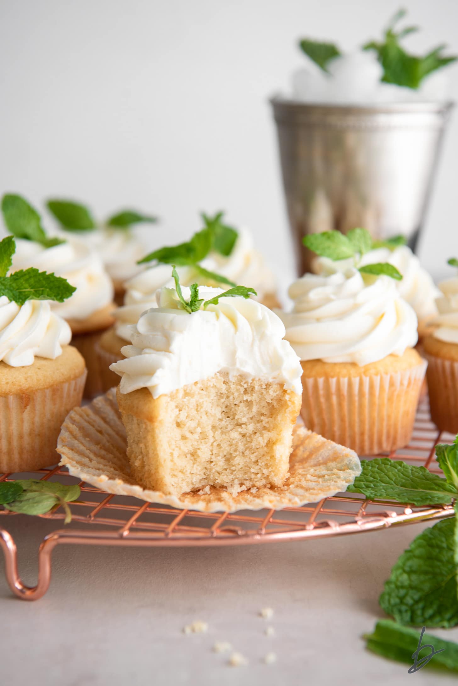mint julep cupcake with a bite on an open paper liner on wire cooling rack with more cupcakes and mint leaves