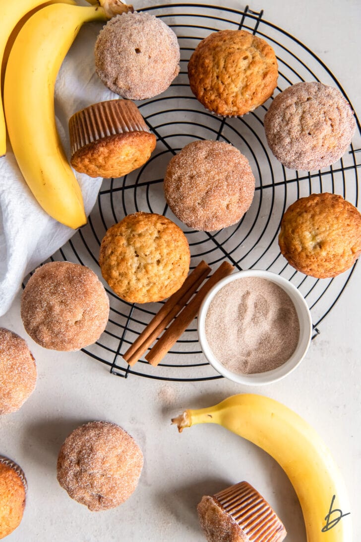 banana cinnamon muffins on a round wire cooling rack next to whole bananas, cinnamon sticks and bowl of cinnamon sugar