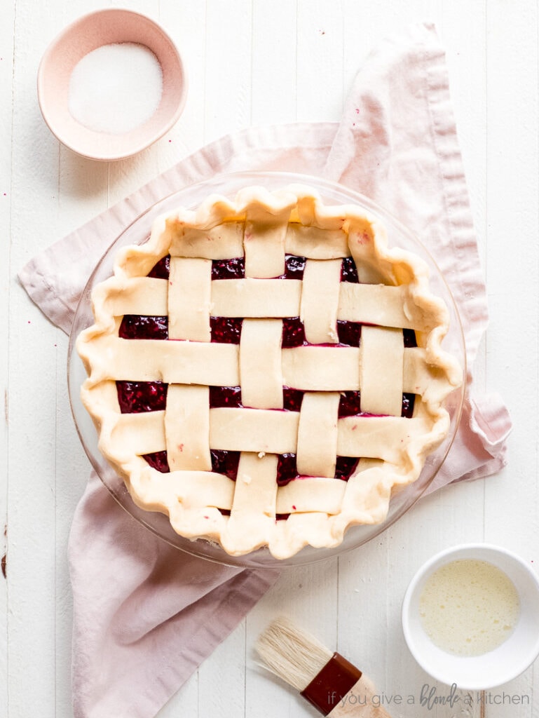 unbaked lattice pie crust with crimped edges. Small pink bowl of sugar. Small white bowl with beaten egg white. Pastry brush.