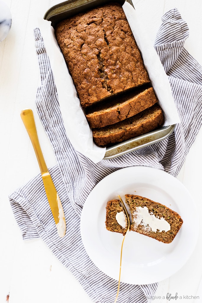 zucchini bread with loaf pan, gold knife, buttered slice, cloth and round white plate