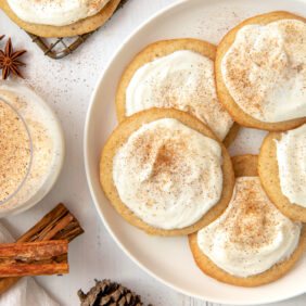 Frosted eggnog cookies on white round plate next to glass of eggnog, small pinecone and cinnamon sticks.