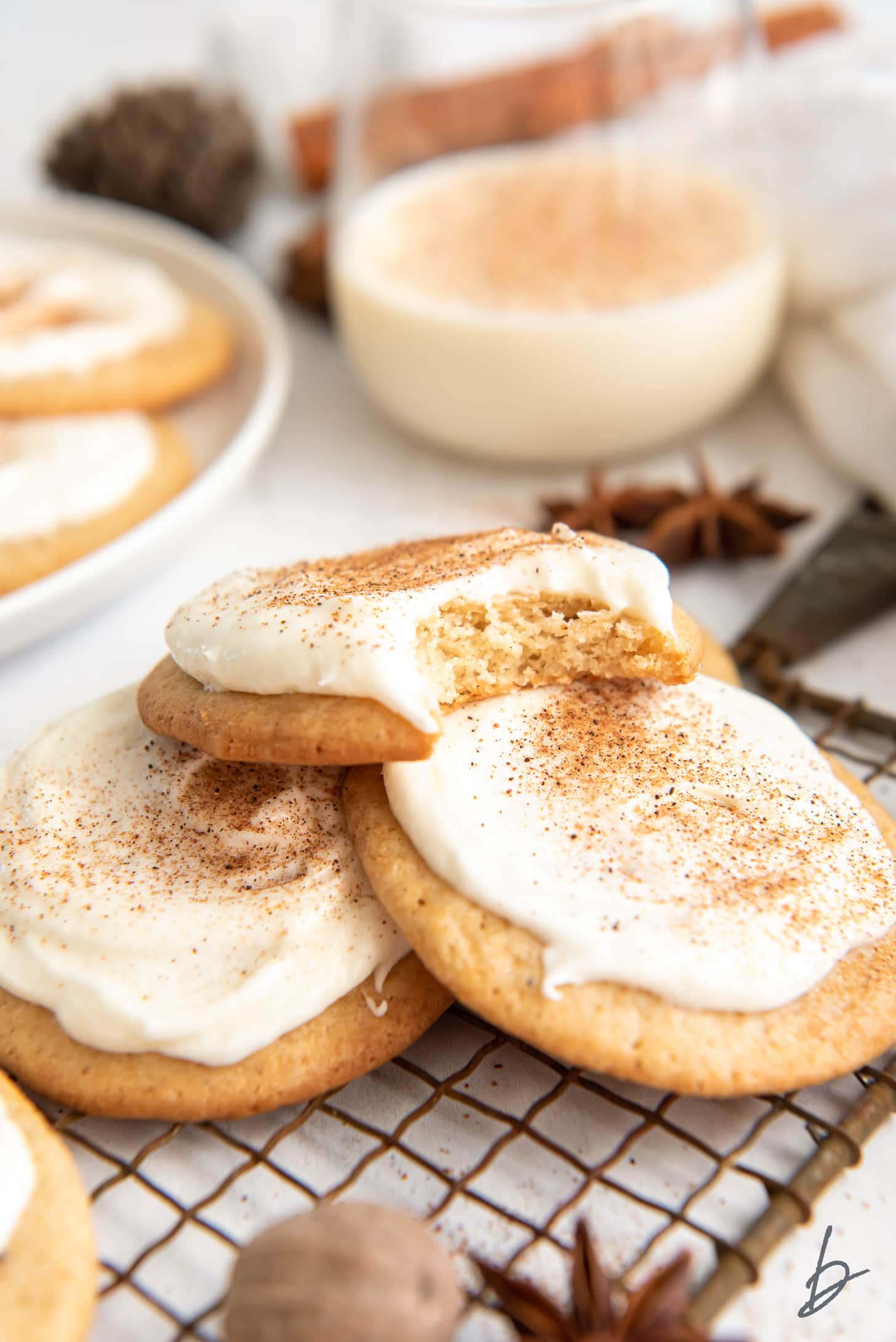 Frosted eggnog cookie with a bite on top of two more cookies on wire cooling rack.