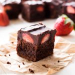 chocolate covered strawberry brownies on parchment paper with strawberries in the background and layer of chocolate ganache on top of fudgy brownie