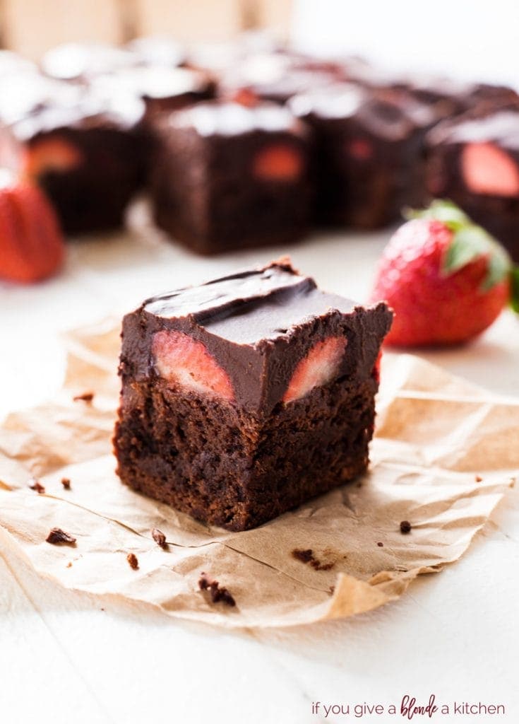 chocolate covered strawberry brownies on parchment paper with strawberries in the background and layer of chocolate ganache on top of fudgy brownie