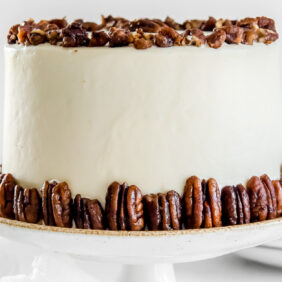 round maple cake with cream cheese frosting and candied pecans on a white cake plate