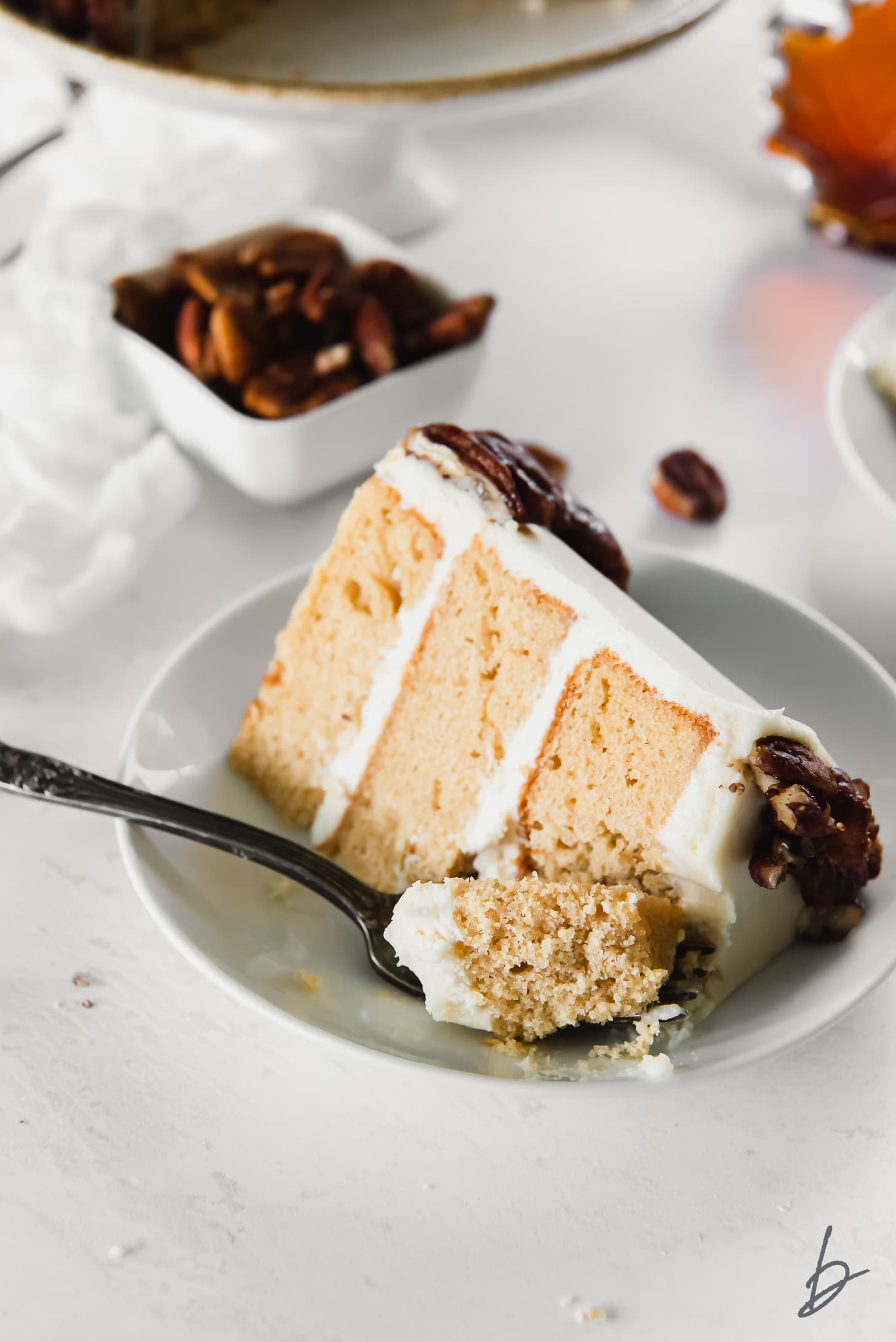 fork taking a bite of layered maple cake slice with cream cheese frosting on a plate