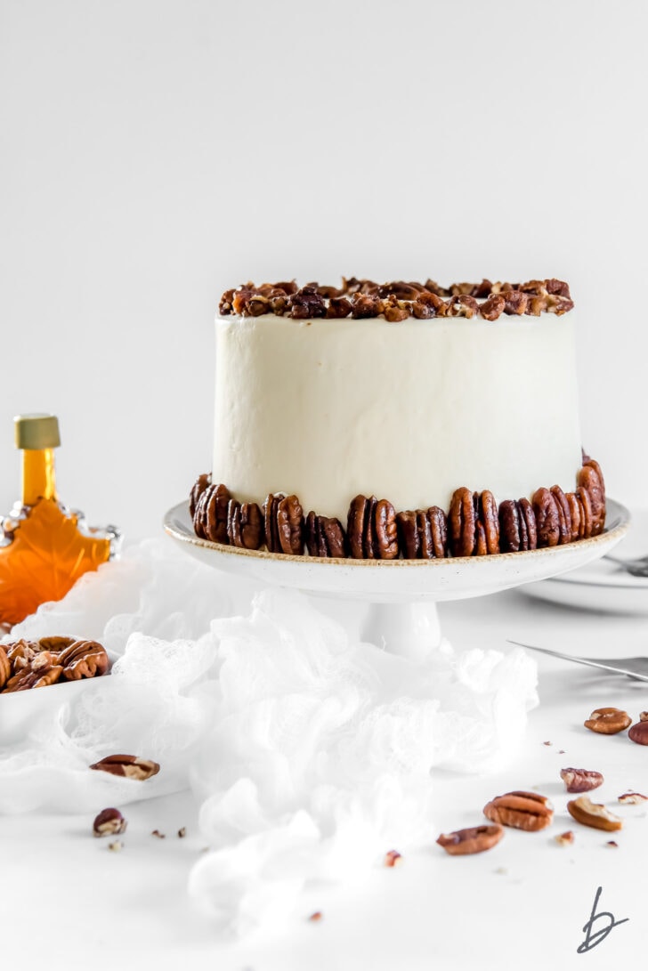 round maple cake with cream cheese frosting and candied pecans on white cake plate