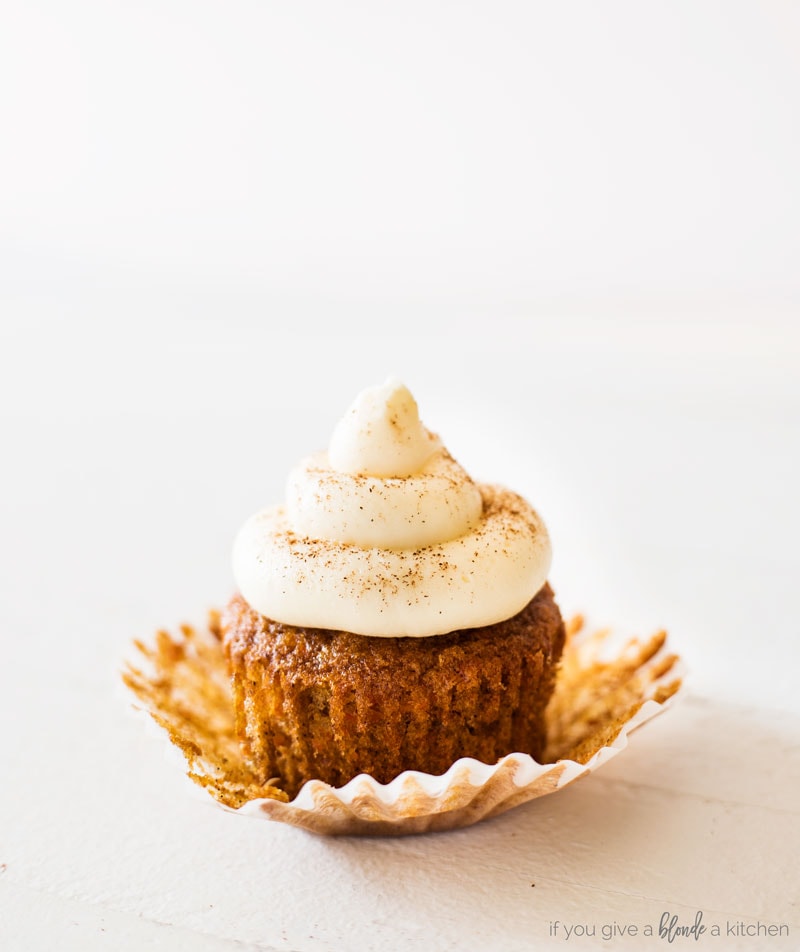 Carrot cake cupcakes with cream cheese frosting. Cupcake sitting on top of open paper liner. Frosting dusted with cinnamon