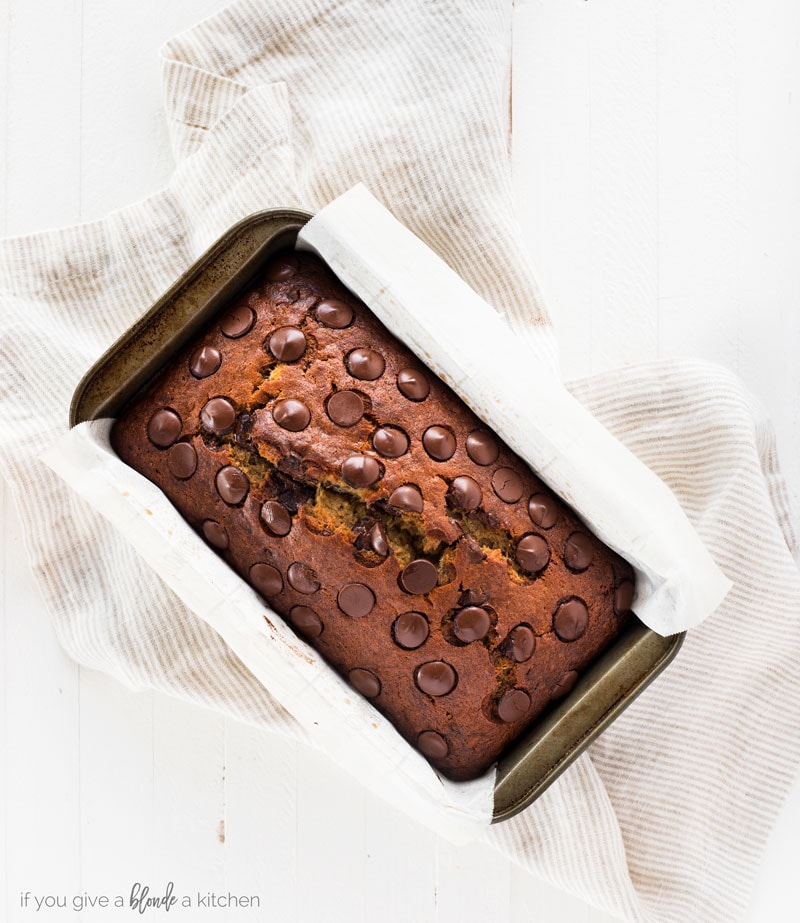Chocolate chip banana bread recipe bread in loaf pan with parchment paper and thin striped kitchen towel on white background