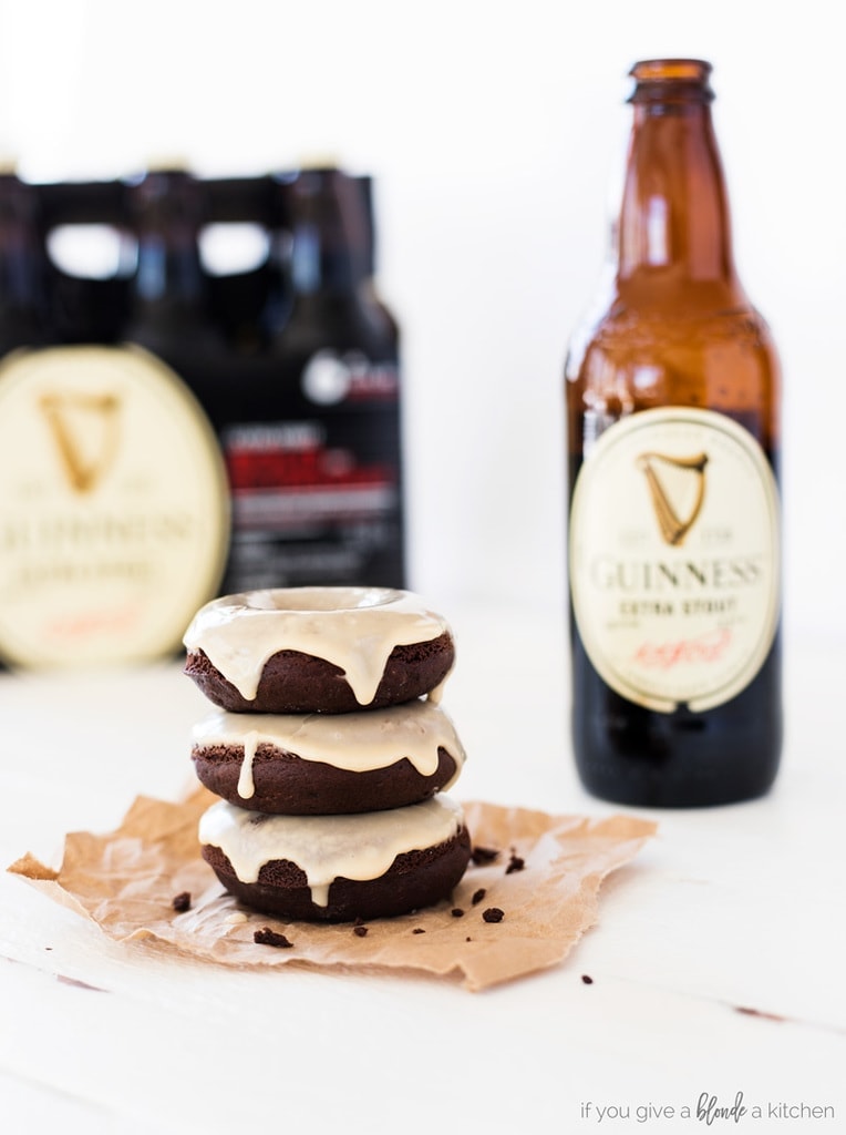 guinness chocolate donuts with bottle of stout beer, stack of three glazed chocolate donuts on brown paper with white wood table