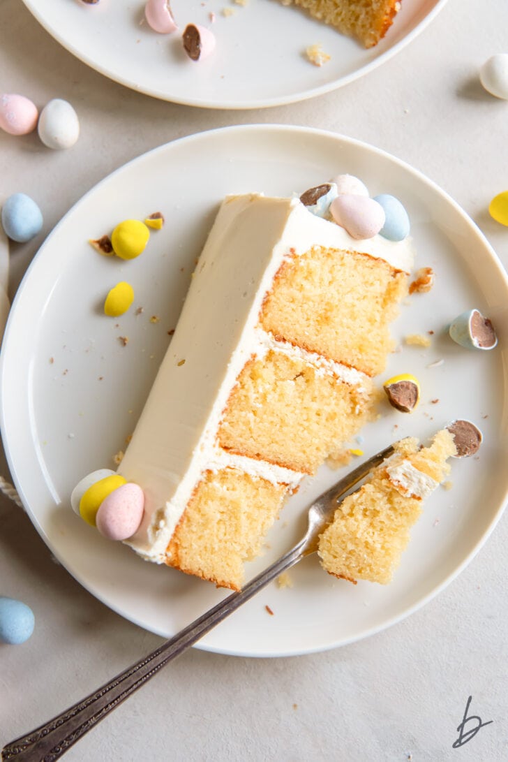 Cadbury mini egg cake slice with a few pastel chocolate eggs on white plate and fork taking a bite of cake