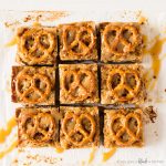 salted caramel pretzel blondies. square blondies with mini pretzel twists on top. symmetry with white background and drizzle of caramel