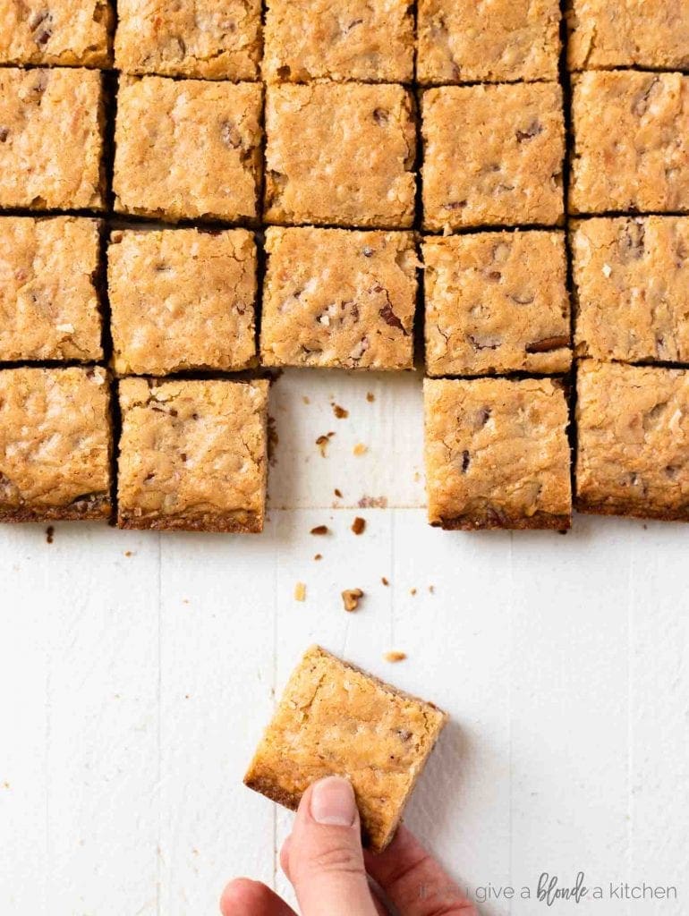 Pecan coconut blondies cut into squares with hand taking one from the symmetrical bars