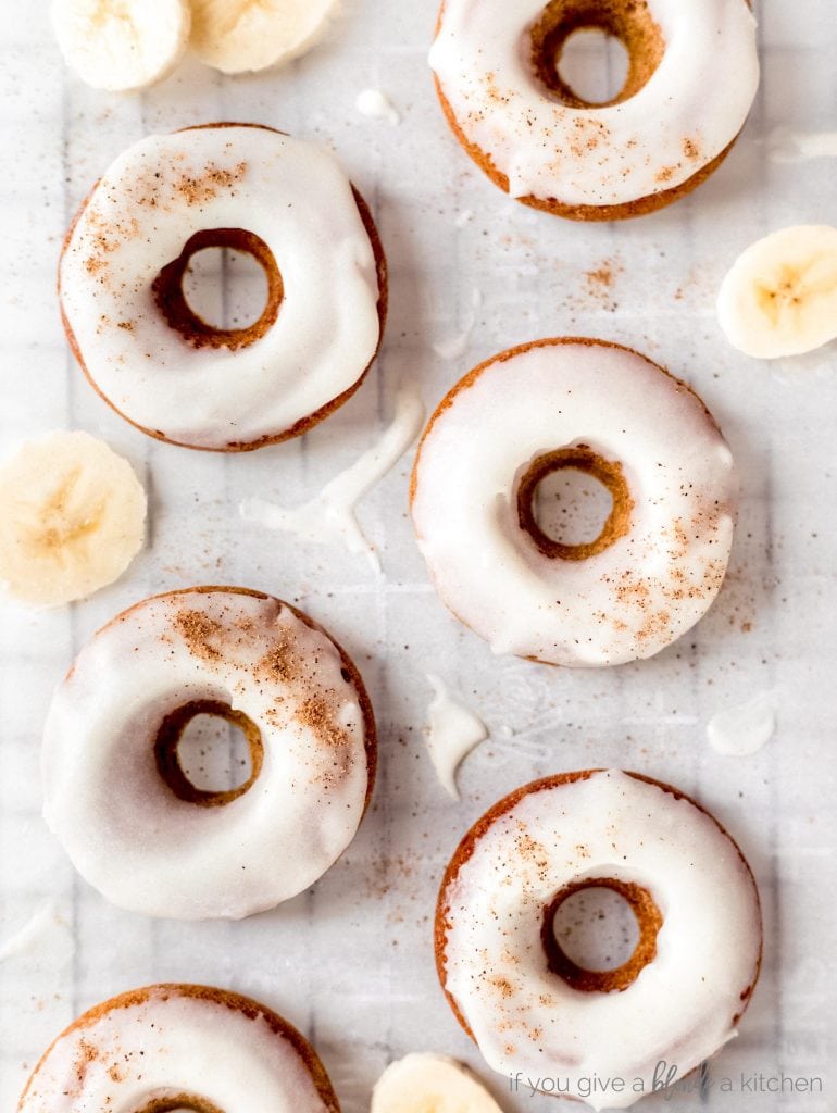 brown butter banana donuts with icing and sprinkle of nutmeg laid flat on parchment paper with banana slices