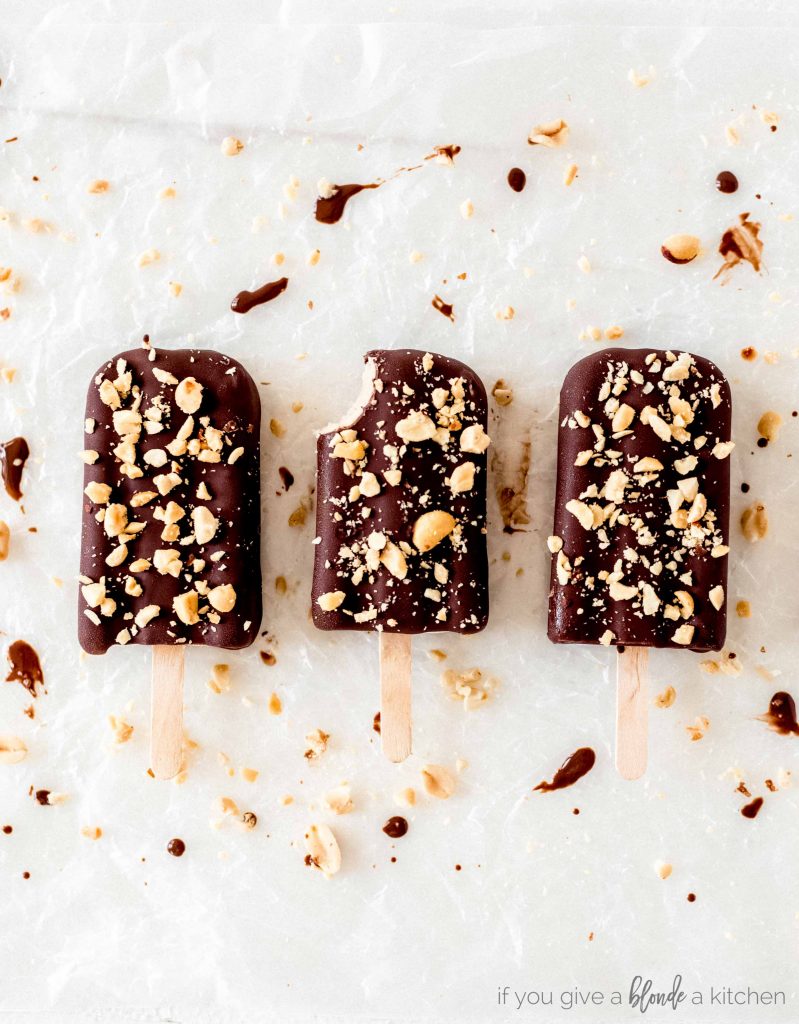 yogurt banana peanut butter popsicles with bite dipped in chocolate with crushed peanuts on parchment paper