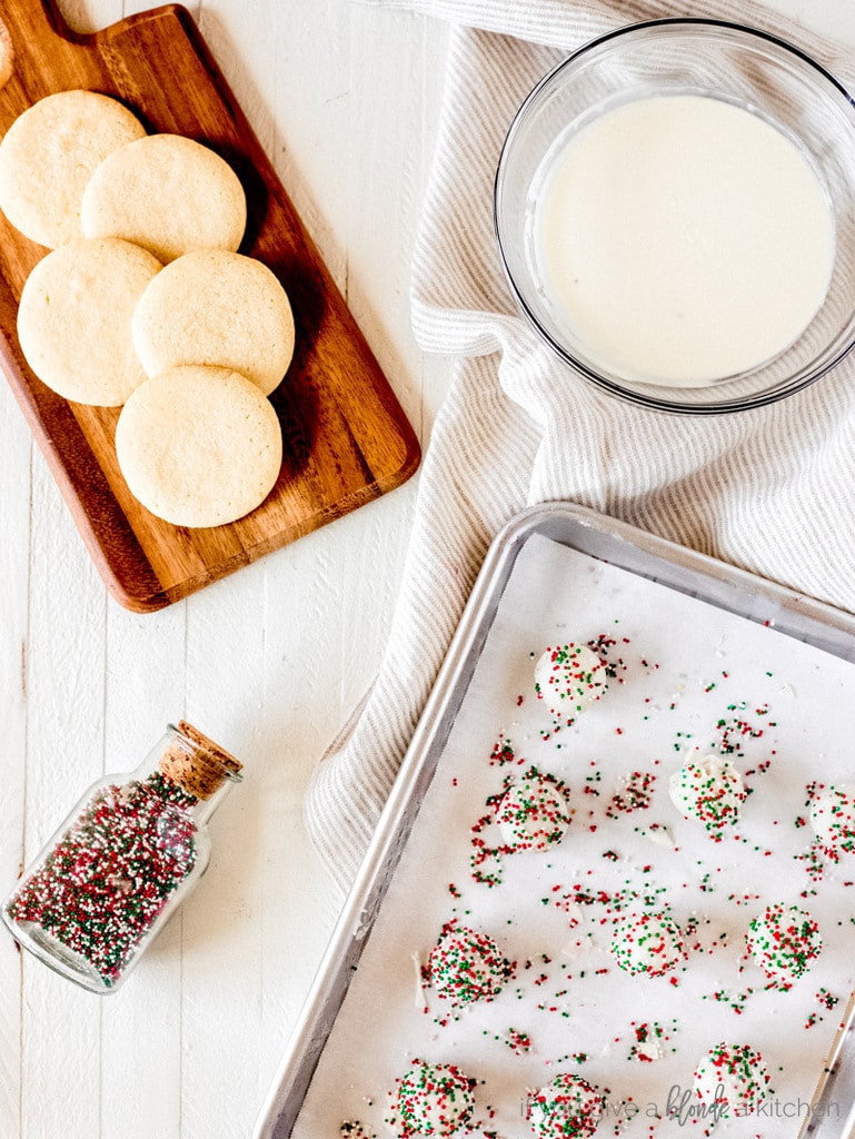 sugar cookie truffles on baing sheet, jat of red and green sprinkles, melted white chocolate and baked sugar cookies