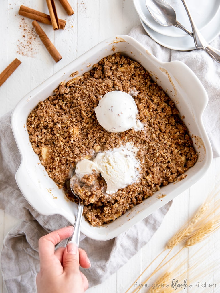 hand holding spoon taking scoop of apple crisp with vanilla ice cream out of baking dish
