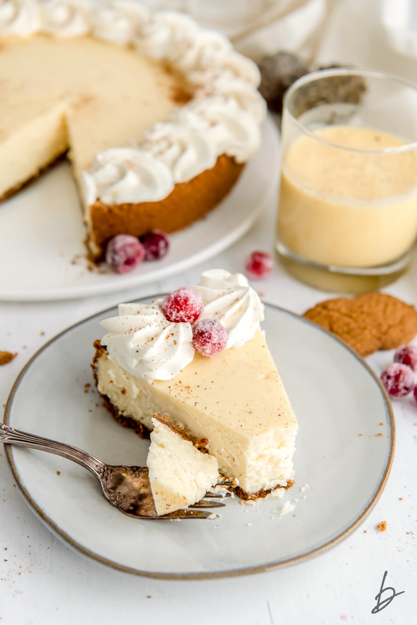 fork taking bite of eggnog cheesecake slice garnished with whipped cream and sugared cranberries.