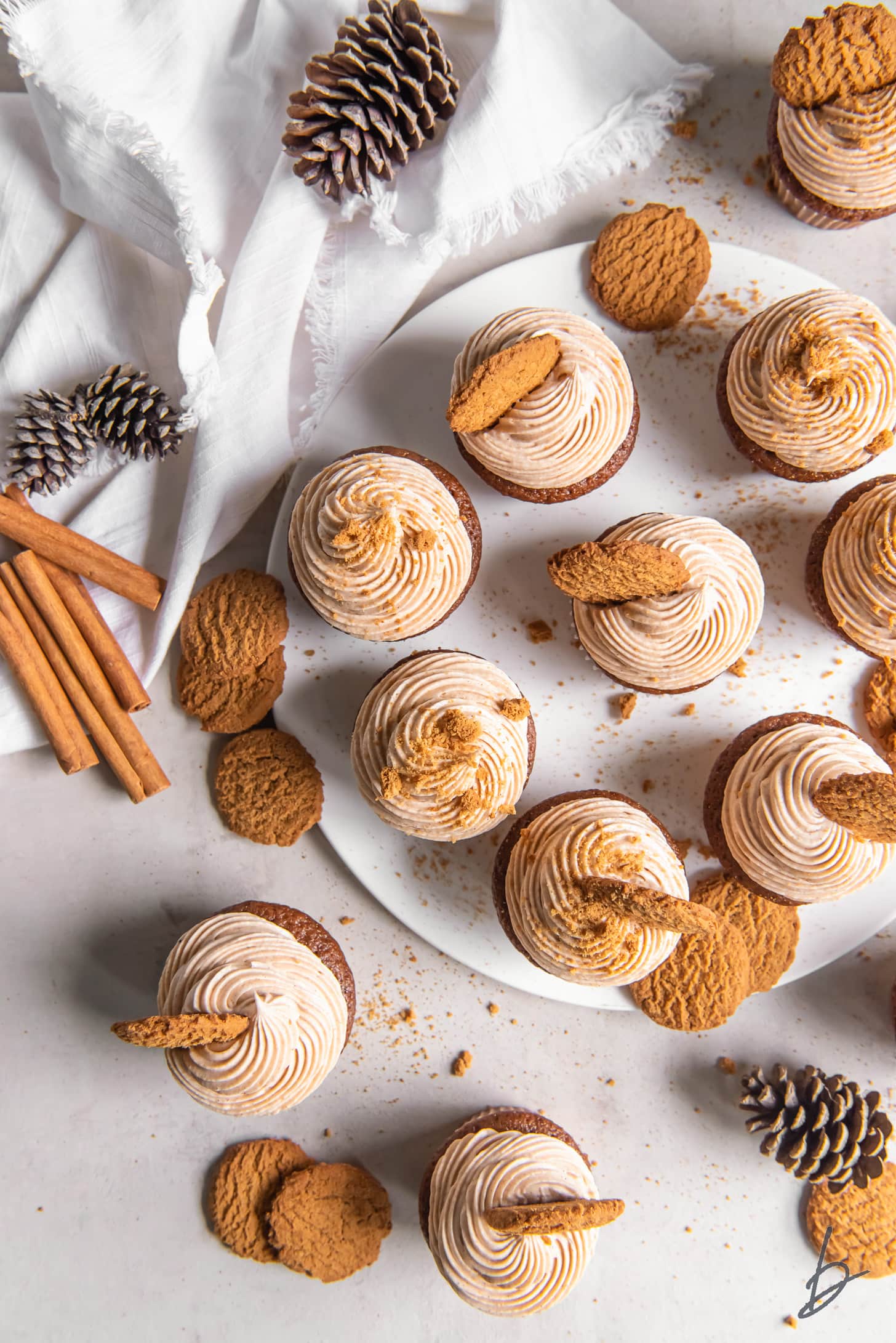 plate of gingerbread cupcakes with frosting and cookies next to pine cones and cinnamon sticks.